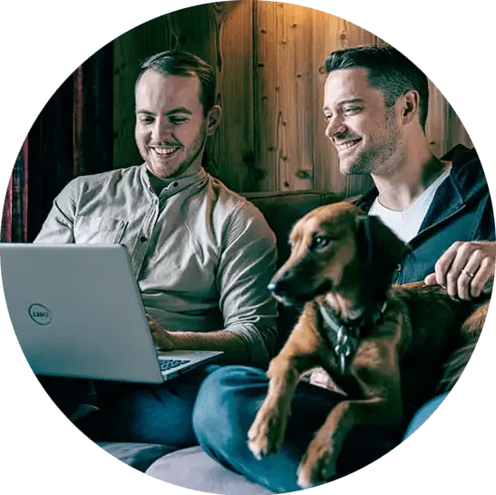 Zwei lächelnde Mitarbeiter schauen auf einen Laptop & haben einen Hund auf dem Schoß
